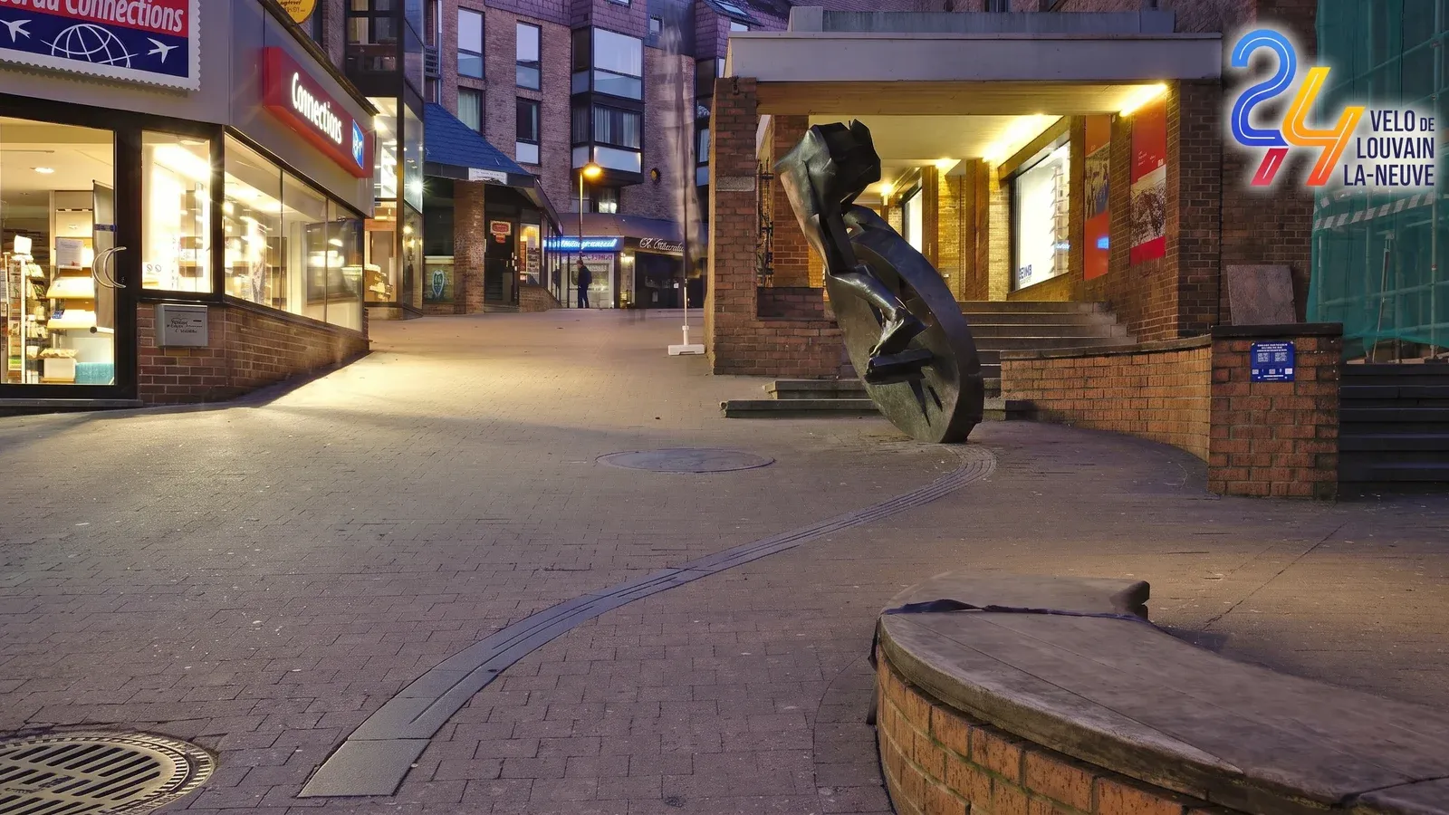 Rue piétonne de Louvain-la-Neuve à la tombée du jour, pavés éclairés et sculpture en forme de roue, avec le logo des 24h vélo de Louvain-la-Neuve