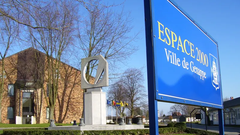 Le panneau bleu « Espace 2000, Ville de Genappe » devant le bâtiment communal en briques et une sculpture, sous un ciel bleu