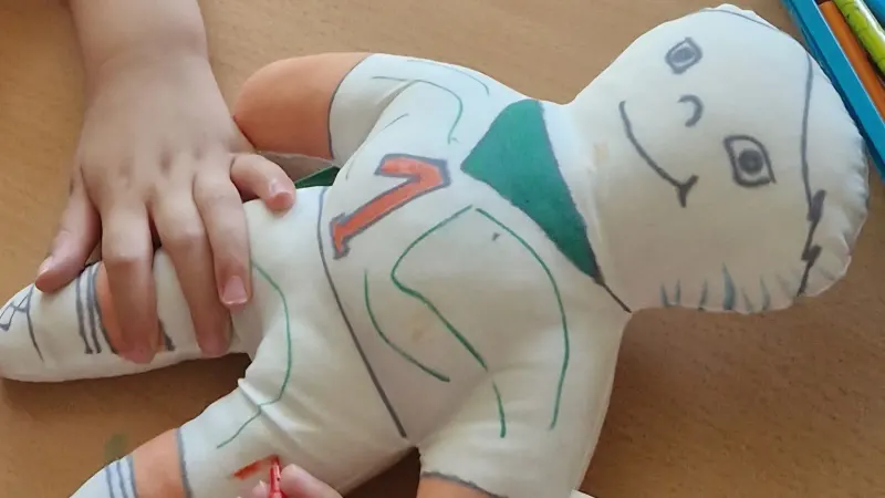 Des mains d’enfant posées sur une poupée en tissu blanc dessinée au feutre en footballeur, maillot numéro 7, sur une table avec des feutres
