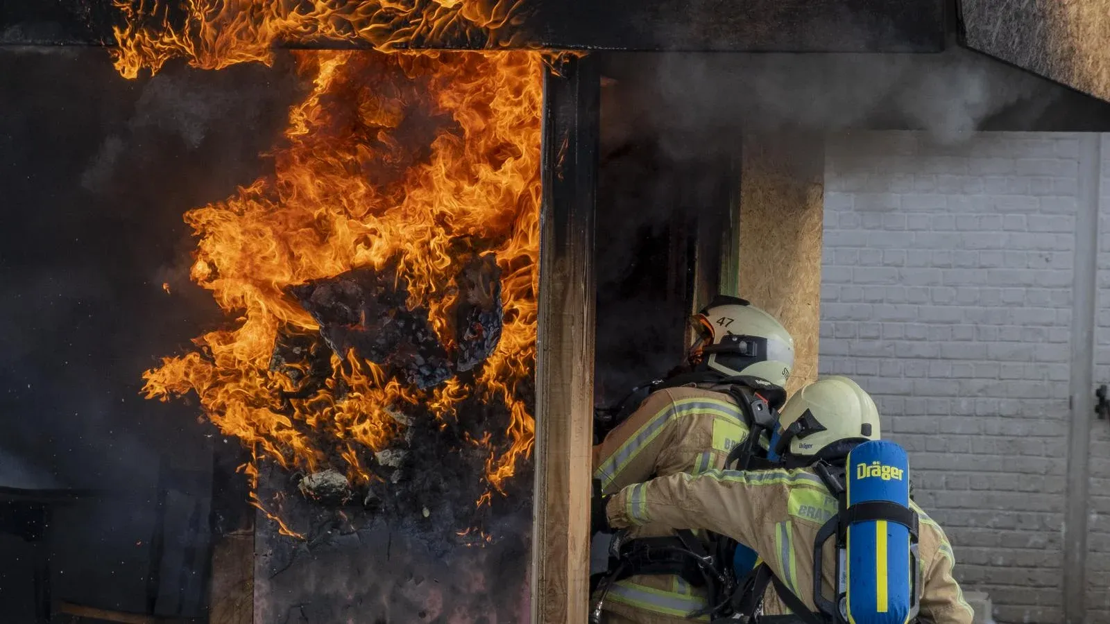 Deux sapeurs-pompiers casqués, de dos, avec leurs bouteilles d'air, avancent vers une porte d'où sortent des flammes, lors d'un exercice