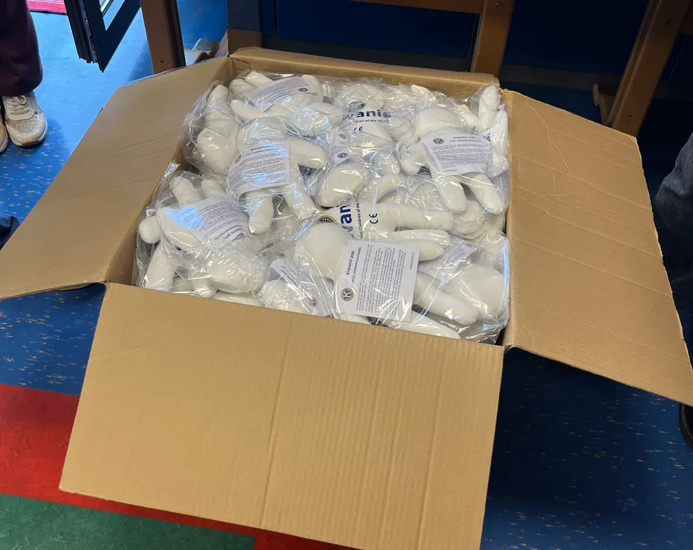 Un grand carton ouvert, rempli de poupées blanches emballées sous plastique avec leur notice, posé au sol devant une table du service de pédiatrie