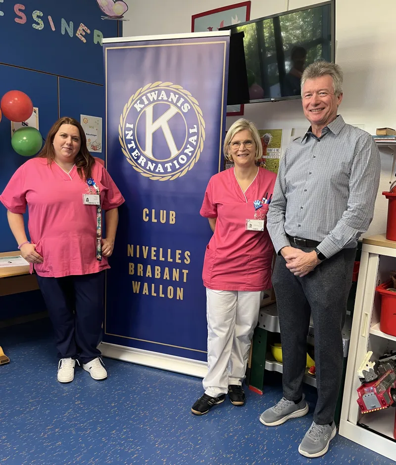 Deux membres de l'équipe de pédiatrie en tenue rose et un membre du Kiwanis posent devant la bannière du Kiwanis International, club Nivelles Brabant wallon, dans le service de pédiatrie
