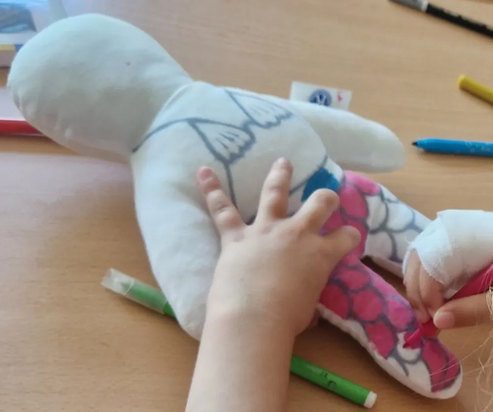 Une main d'enfant tient une poupée Kiwanis décorée en sirène, aux jambes couvertes d'écailles roses, pendant qu'une autre main, bandée, la colorie au feutre